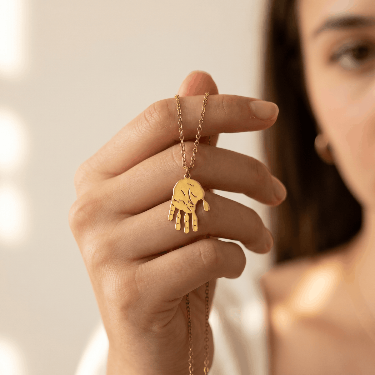 Frau hält goldfarbene Handabdruck-Halskette mit konturgenau ausgeschnittenem Baby-Handabdruck-Anhänger zwischen den Fingern (KI-Visualisierung)