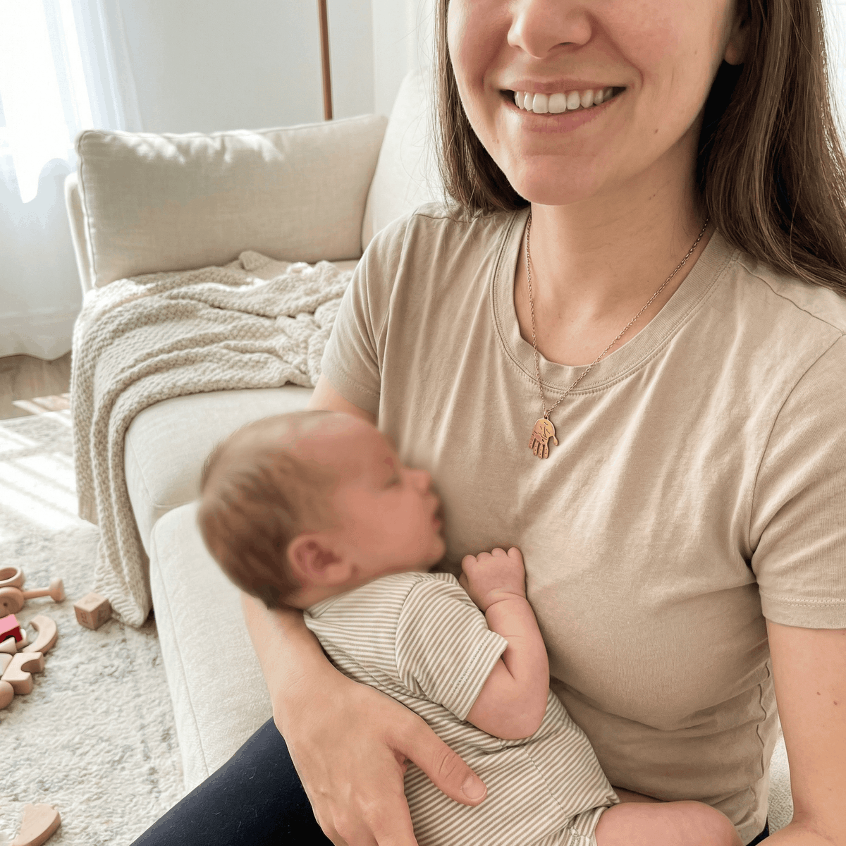 Lächelnde Mutter trägt goldfarbene Handabdruck-Halskette mit konturgenau ausgeschnittenem Baby-Handabdruck und hält ihr Baby (KI-Visualisierung)